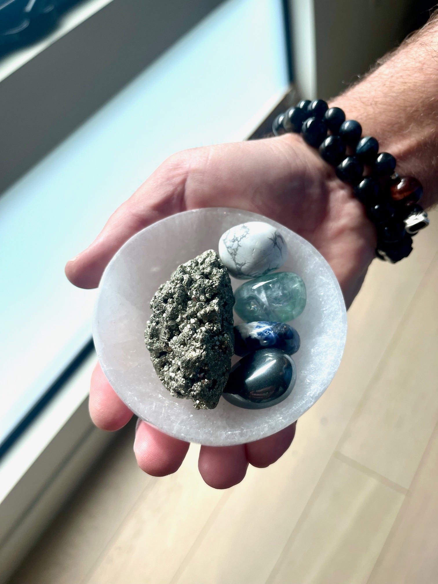 Male hand holding a selenite charging bowl with healing crystal set of pyrite, hematite, green fluorite, white howlite and sodalite.
