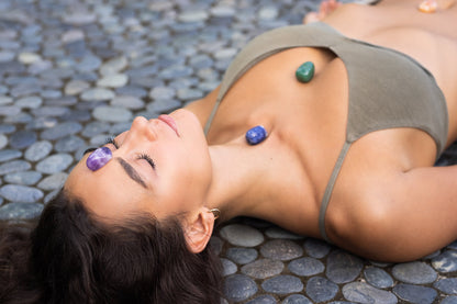 dark haired woman laying on stone ground with amethyst crystal on her crown chakra, sodalite stone on her throat chakra and green aventurine on her heart chakra and tigers eye on her solar plexus chakra