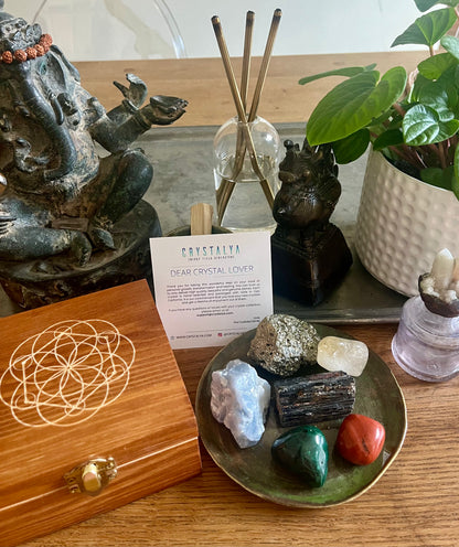 feng shui healing crystal set on display next to engraved seed of life wooden keepsake box with healing stones next to it on green plate - pyrite, black tourmaline, malachite, red jasper, blue calcite and citrine next to ganesh statue and house plant.
