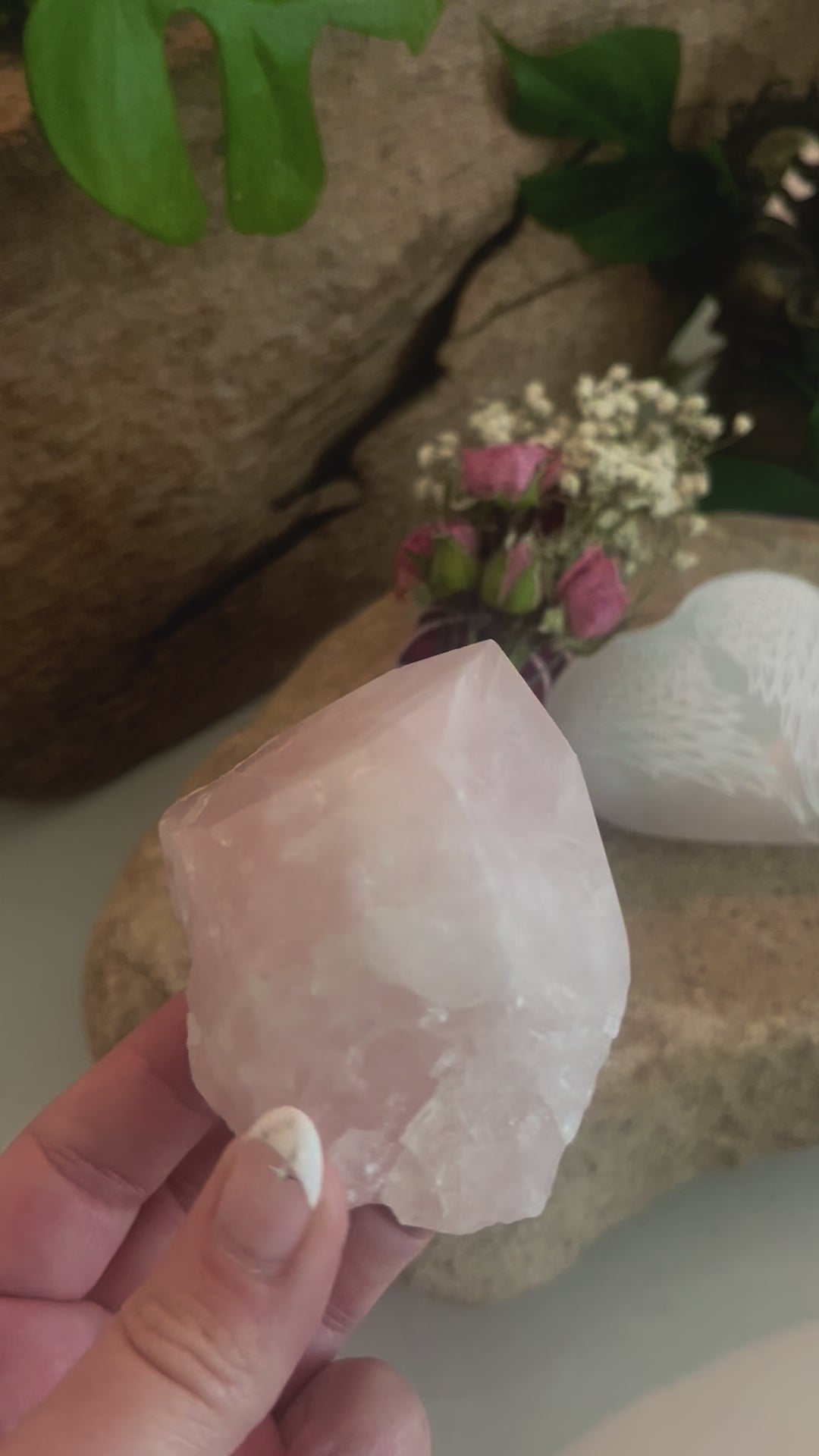 Hand-held Rose quartz crystal being displayed with selenite heart-shaped stone with engraved angel wings and floral rose sage smudge bundle sitting on a stone in the background.