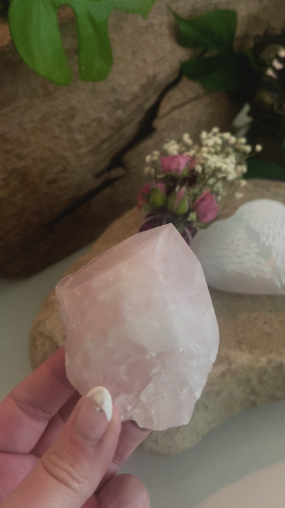 Hand-held Rose quartz crystal being displayed with selenite heart-shaped stone with engraved angel wings and floral rose sage smudge bundle sitting on a stone in the background.